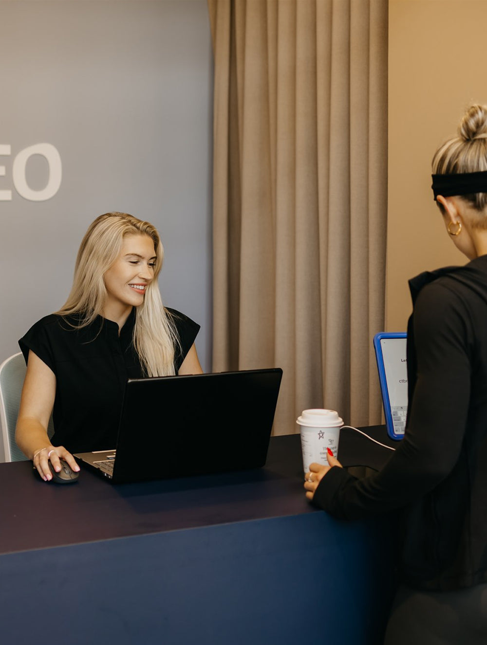 Two women at Cleo front desk with laptops and a coffee cup, engaged in a conversation.