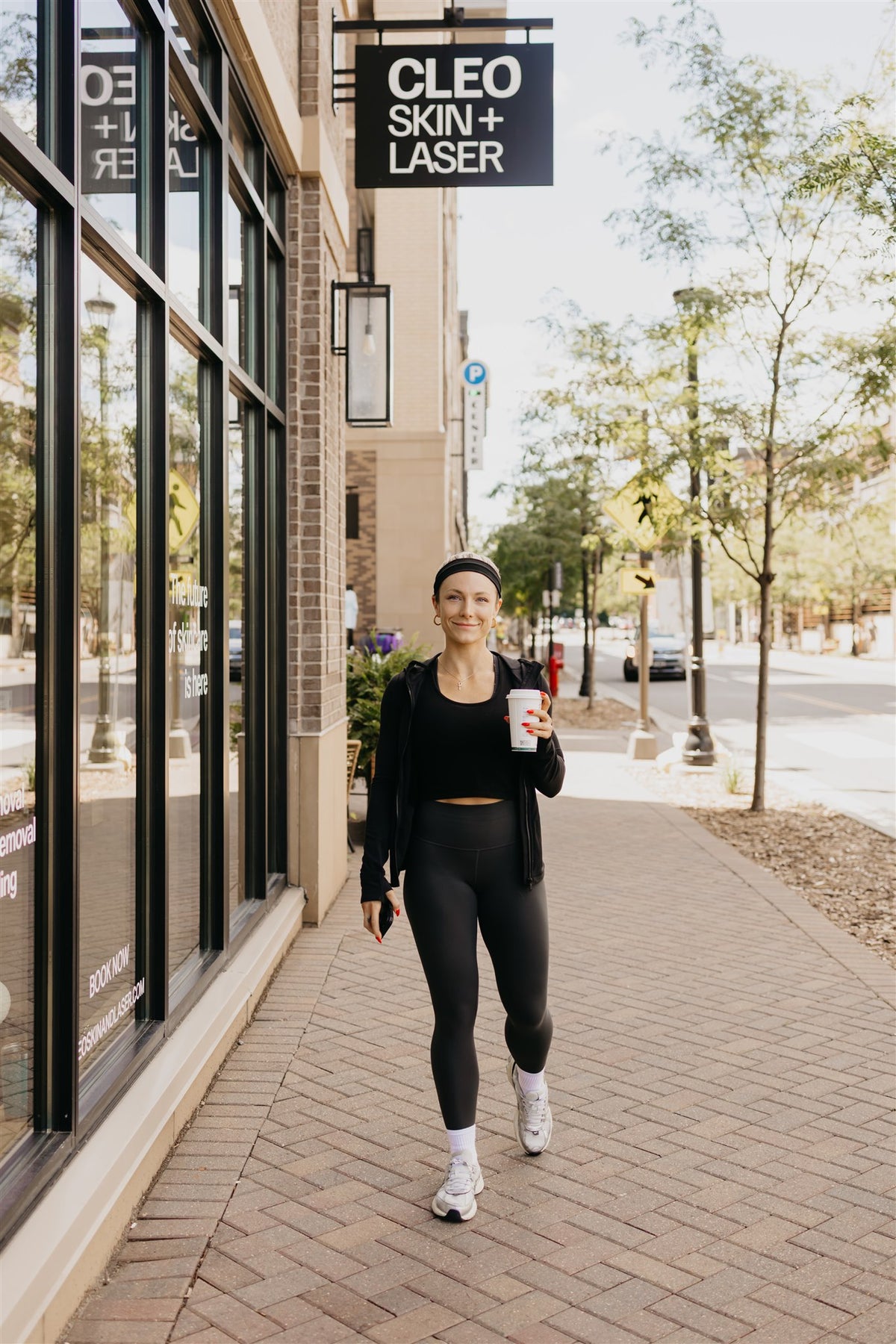 A woman in a black athletic outfit walks on a brick sidewalk in front of a storefront with a sign reading 'CLEO SKIN+ LASER', holding a white coffee cup.