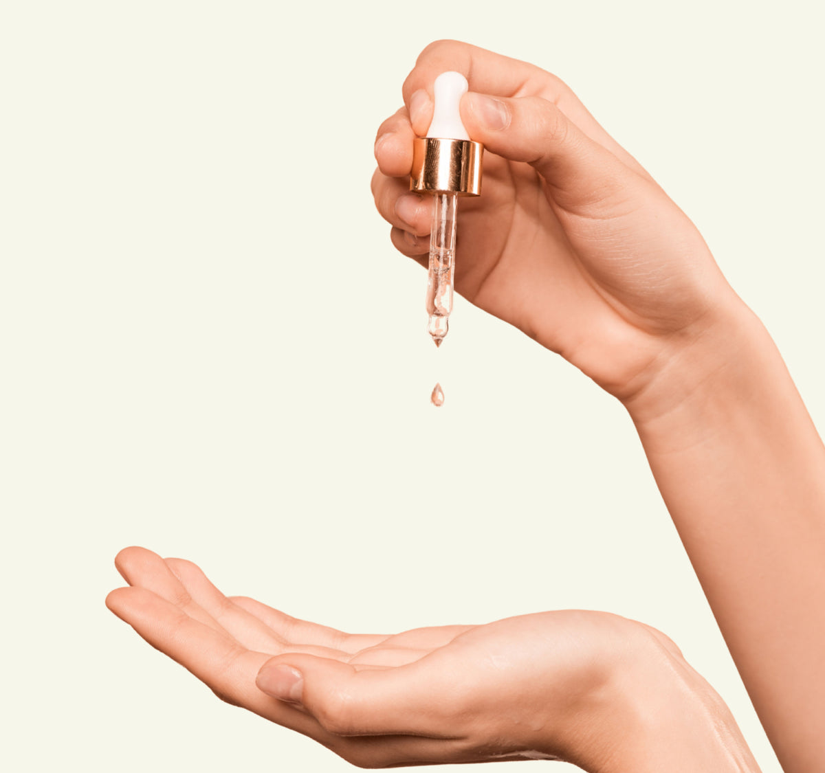 A hand holding a glass dropper bottle with a gold collar and white rubber bulb, with a single drop of pinkish serum falling, while another hand is open below. The background is a solid pale cream color.