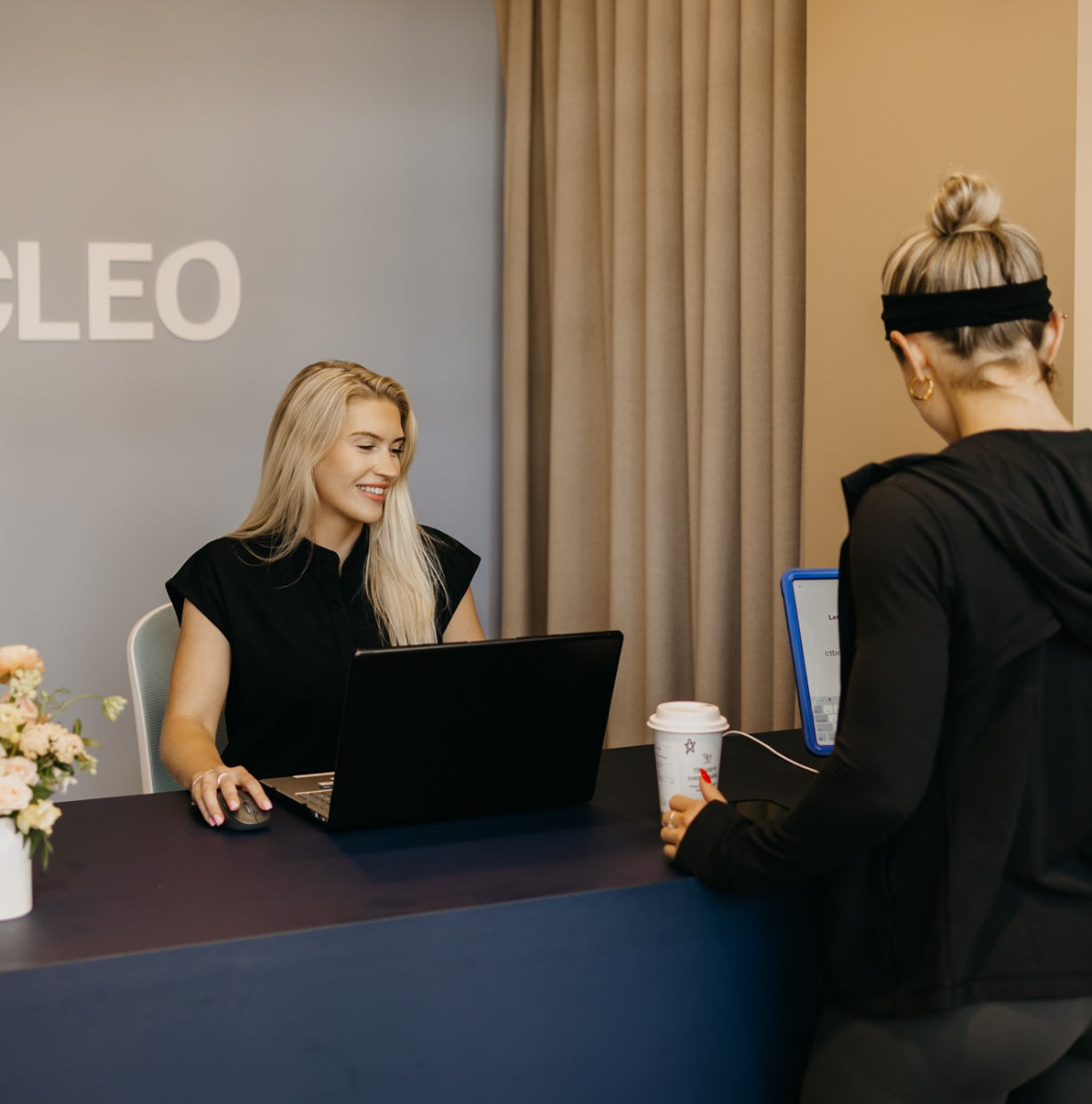 Two women at a desk with a laptop, one using a mouse, in an office setting with 'Cleo' branding.