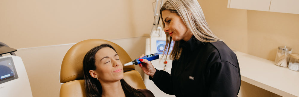A dental or skincare professional in a black uniform applies a blue and silver cosmetic device to a woman's face while she reclines in a tan chair inside a clinic room.
