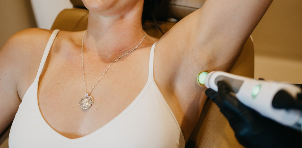 A close-up of a woman's underarm while wearing a white tank top and silver pendant necklace, with a gloved hand holding a handheld device against the armpit.